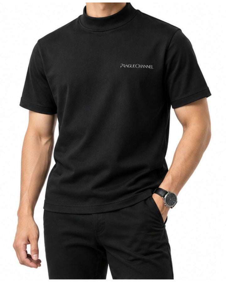 Muž v černém tričku s nízkým stojáčkem a černých kalhotách, ruka v kapse